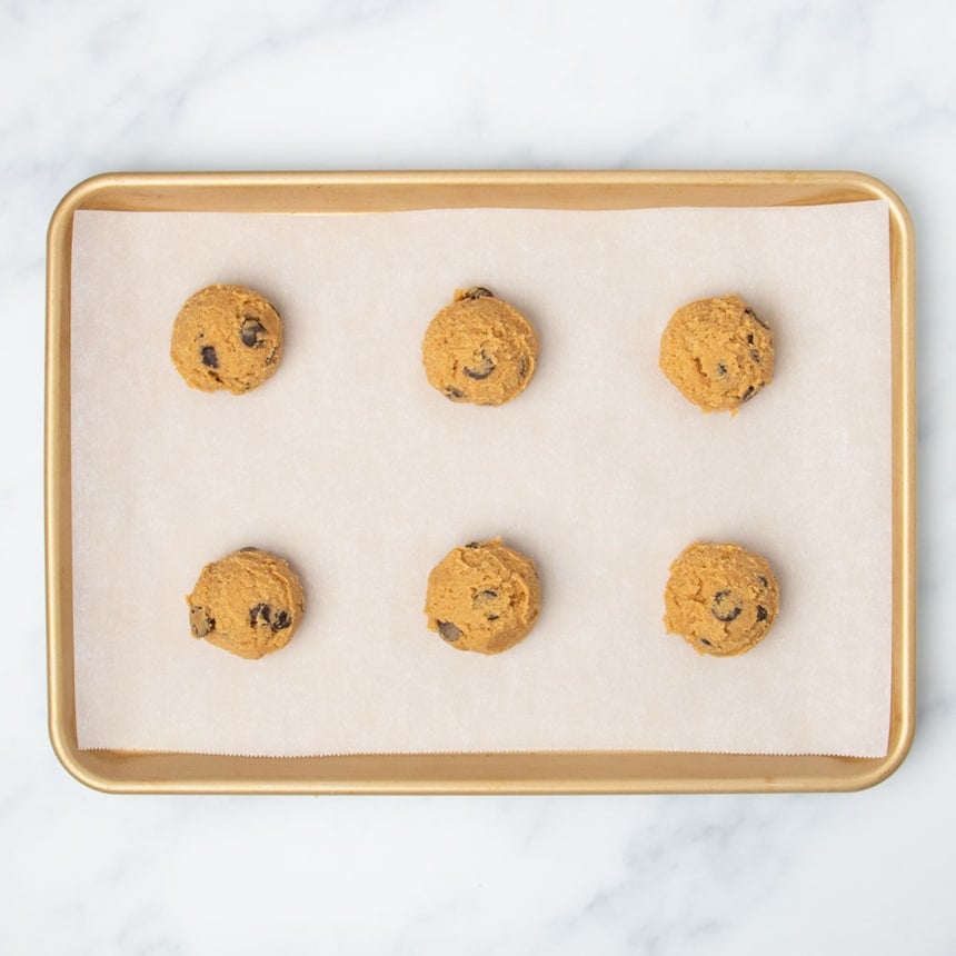 Overhead image of 6 raw rounds of light orange cookie dough with chocolate chips spaced evenly apart on white paper on gold rimmed baking sheet