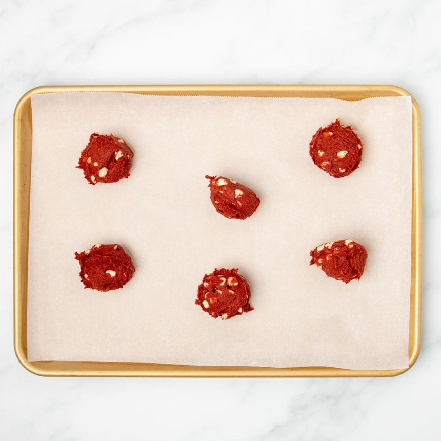 6 irregular mounds of red velvet cookie dough on white paper lined gold baking tray