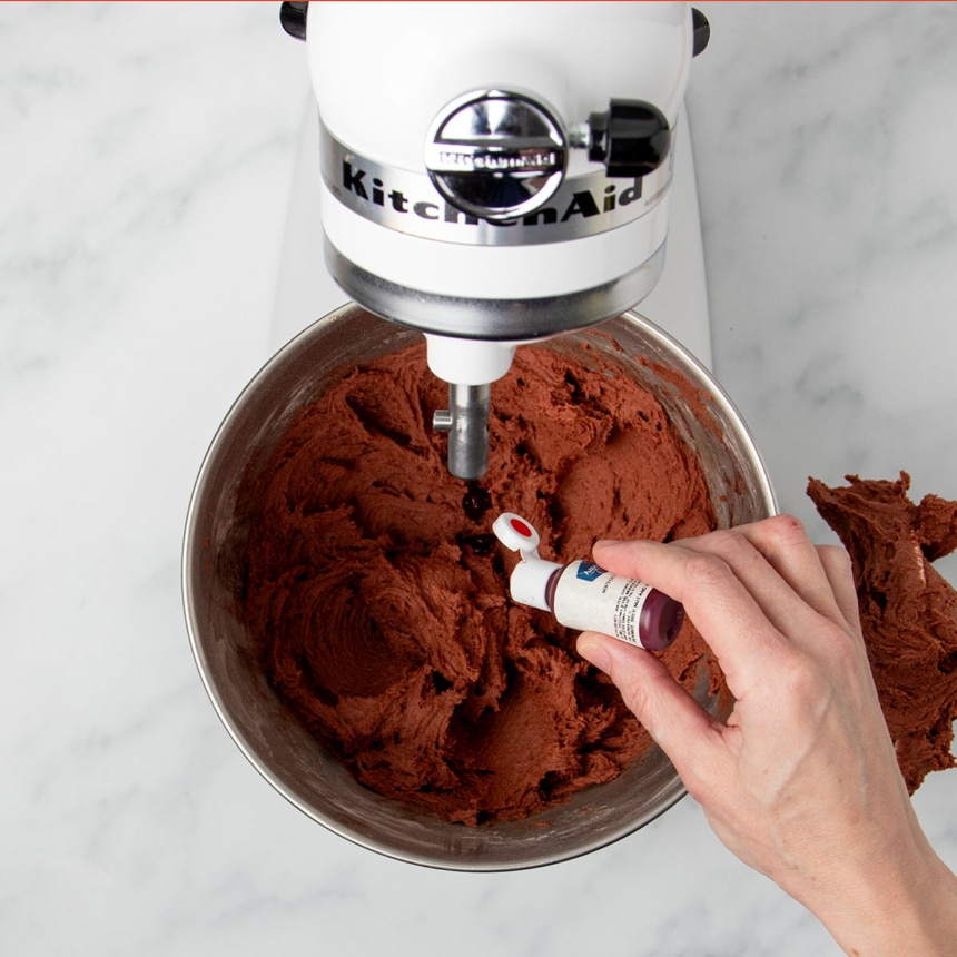 hand squeezing small bottle of red gel food coloring into brown cookie dough in bowl