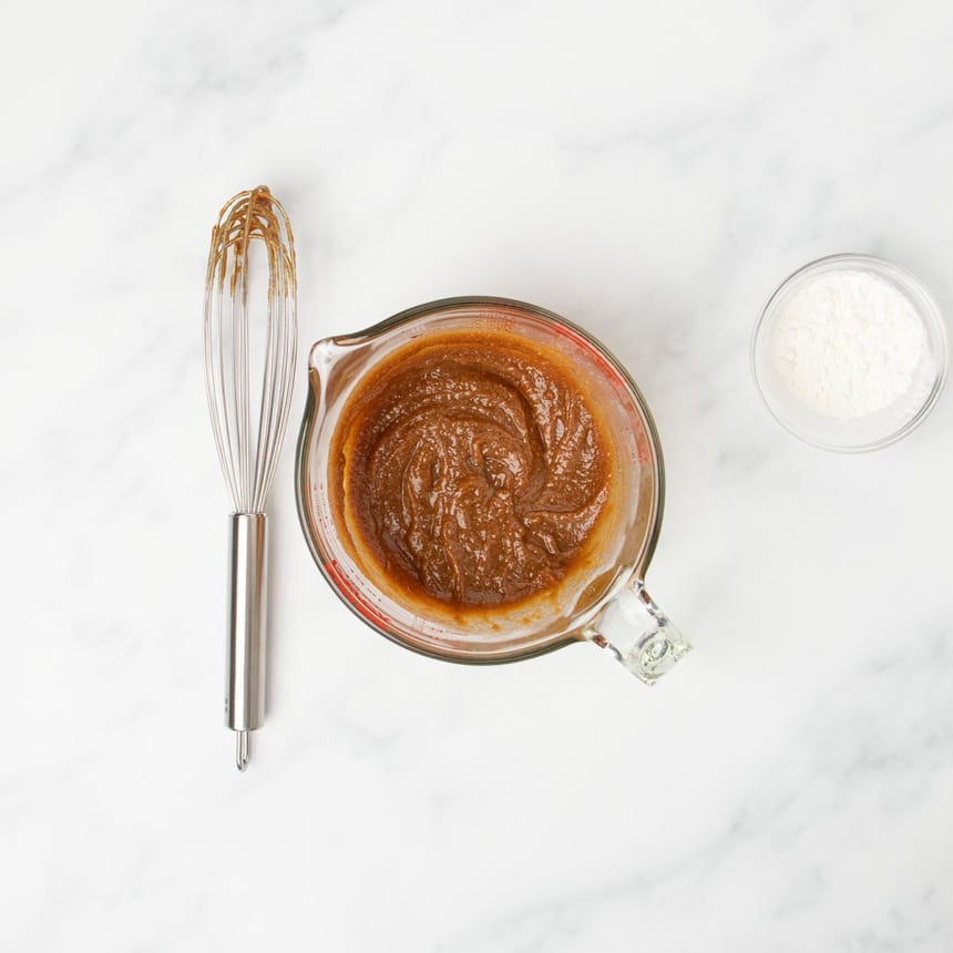 thick brown pasta in same glass measuring cup with wire whisk on marble table next to it