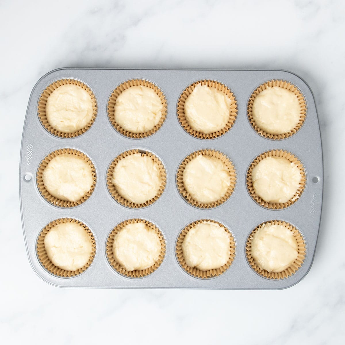 standard 12 cup metal muffin tin with brown muffin liners in wells filled 2/3 of the way full with light yellow raw cupcake batter