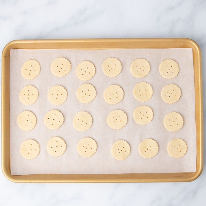 24 rounds of raw gluten free ritz crackers with holes on white paper on gold rimmed baking sheet