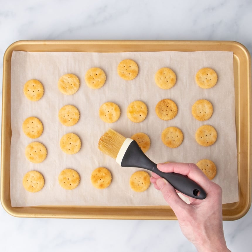 hand holding pastry brush, brushing one of 24 baked light brown ritz crackers with melted butter