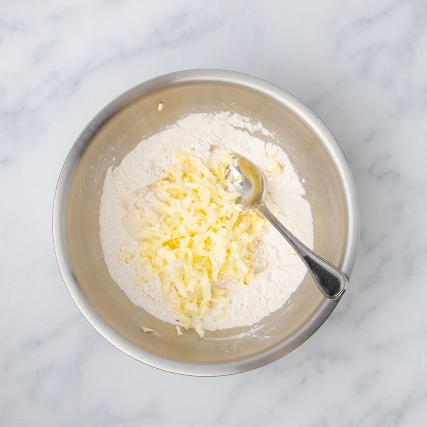 shreds of yellow grated butter on top of white powder in mixing bowl, with metal mixing spoon