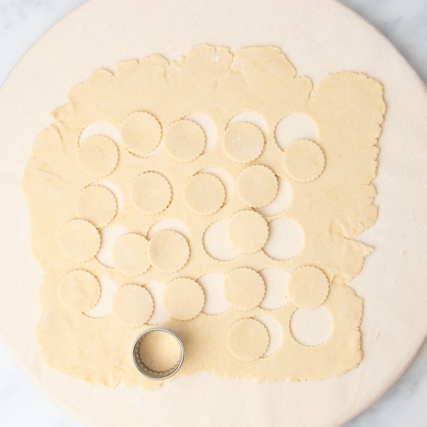 dough rolled out flat on board with scalloped-edge small round metal cookie cutter with lots of round cut out and resting on dough