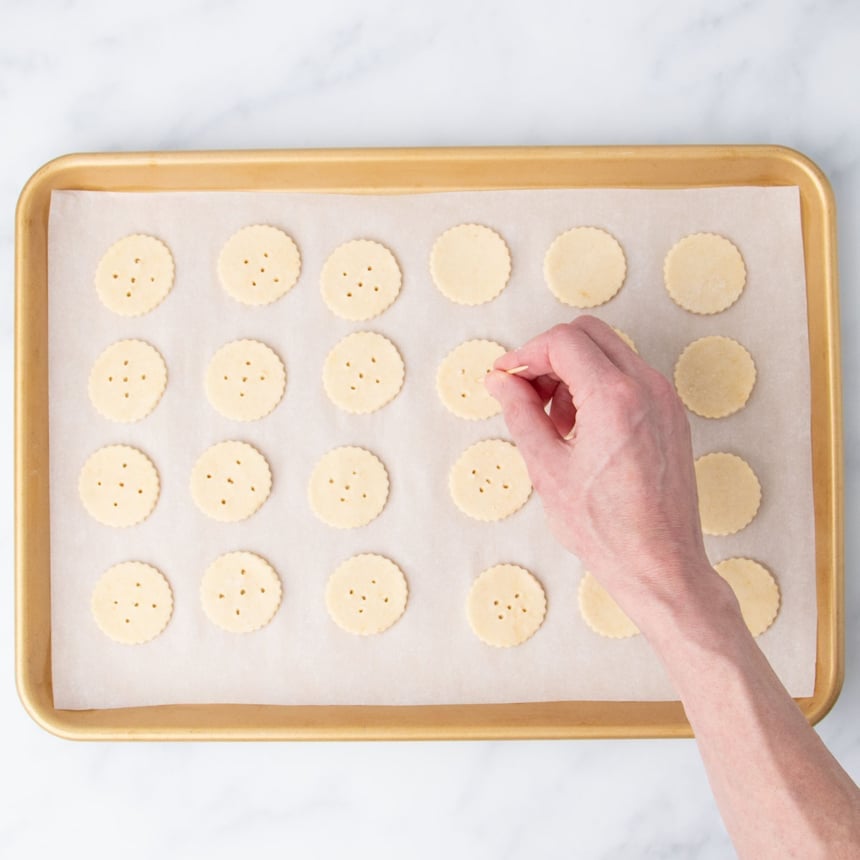 24 raw cracker rounds with 5 holes each with hand holding toothpick pressing holes into rounds