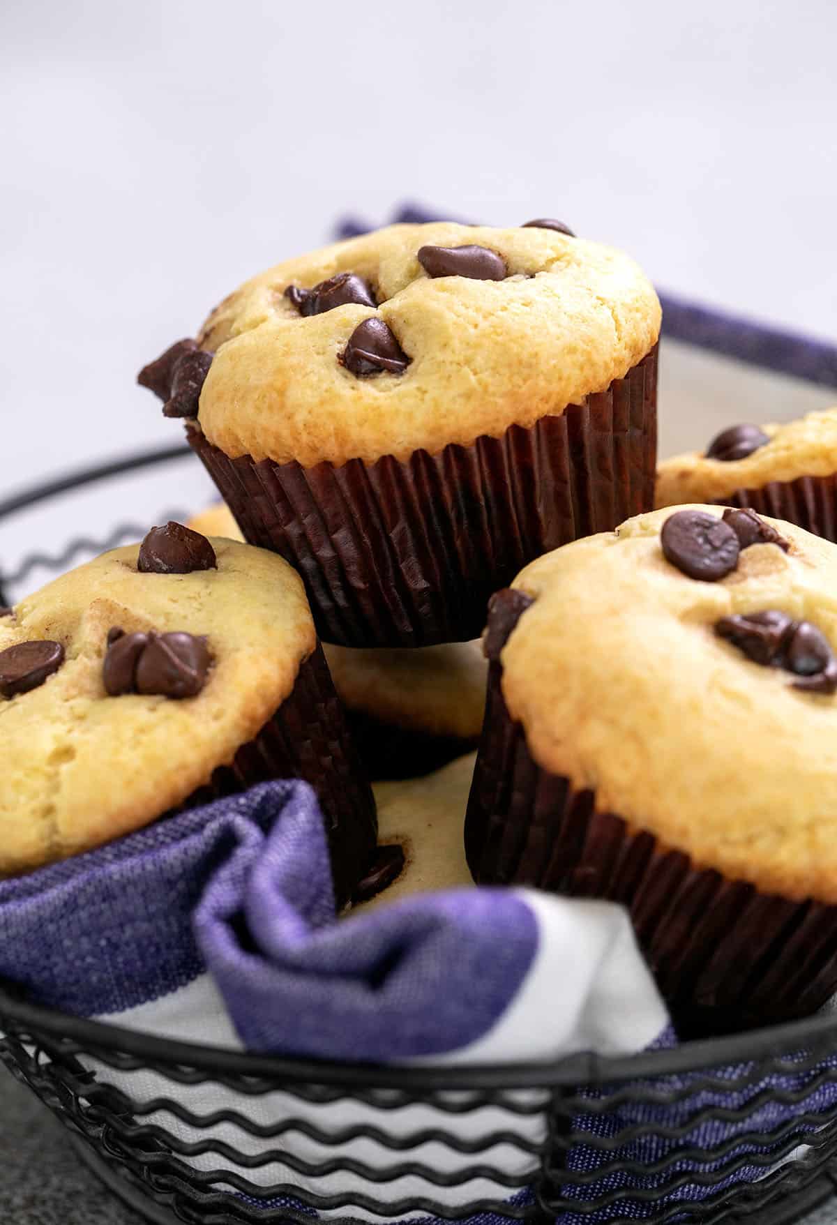 Pile of chocolate chip muffins in brown liners in a wire basket lined with blue and white striped towel