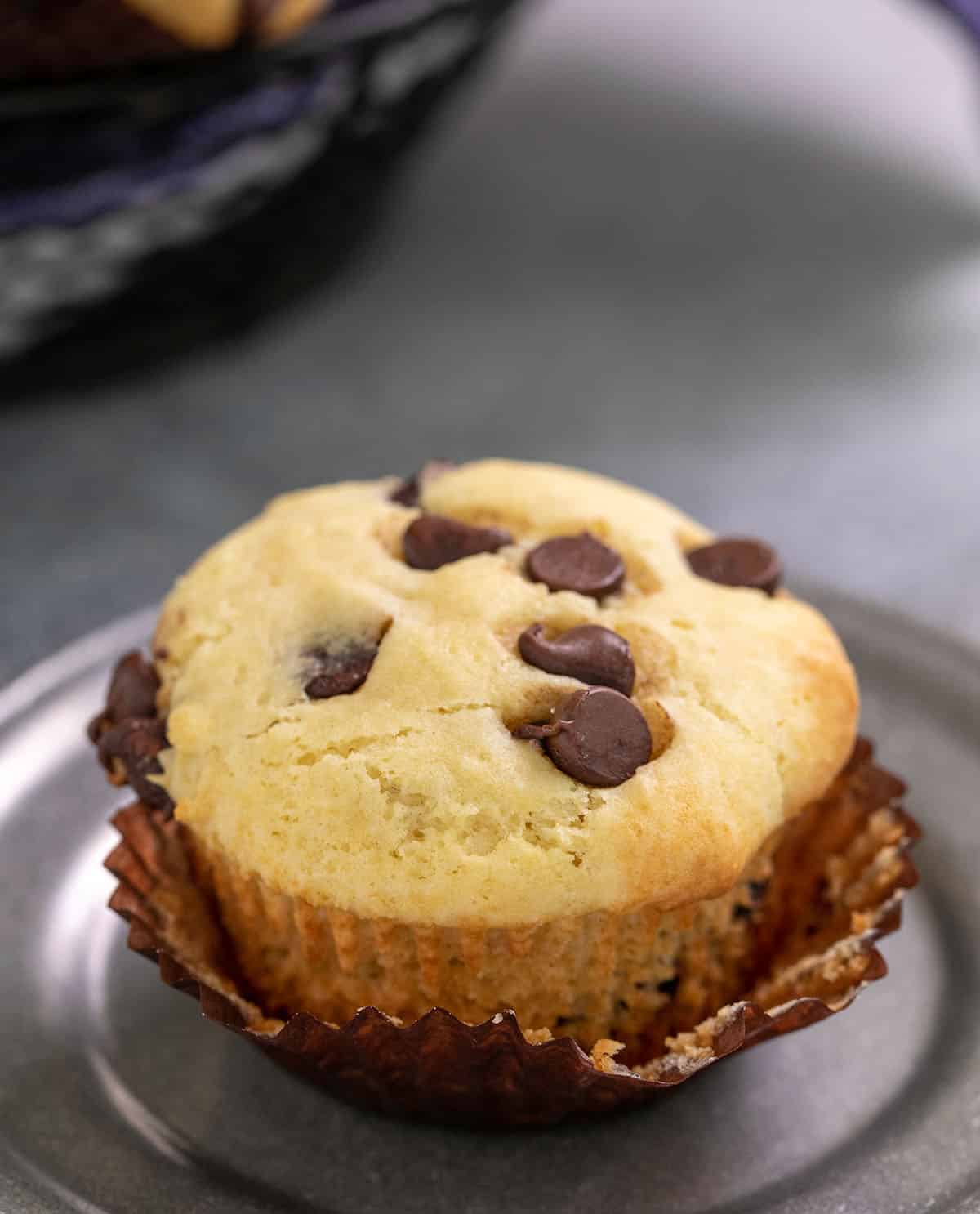 Chocolate chip muffin with muffin liner partially peeled off on a small pewter plate and muffin basket in background