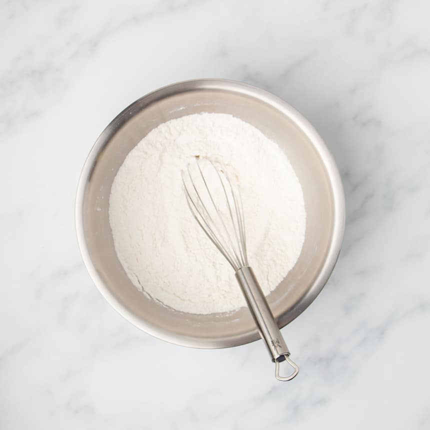 Large round metal mixing bowl with white flour and other white ingredients with large wire whisk