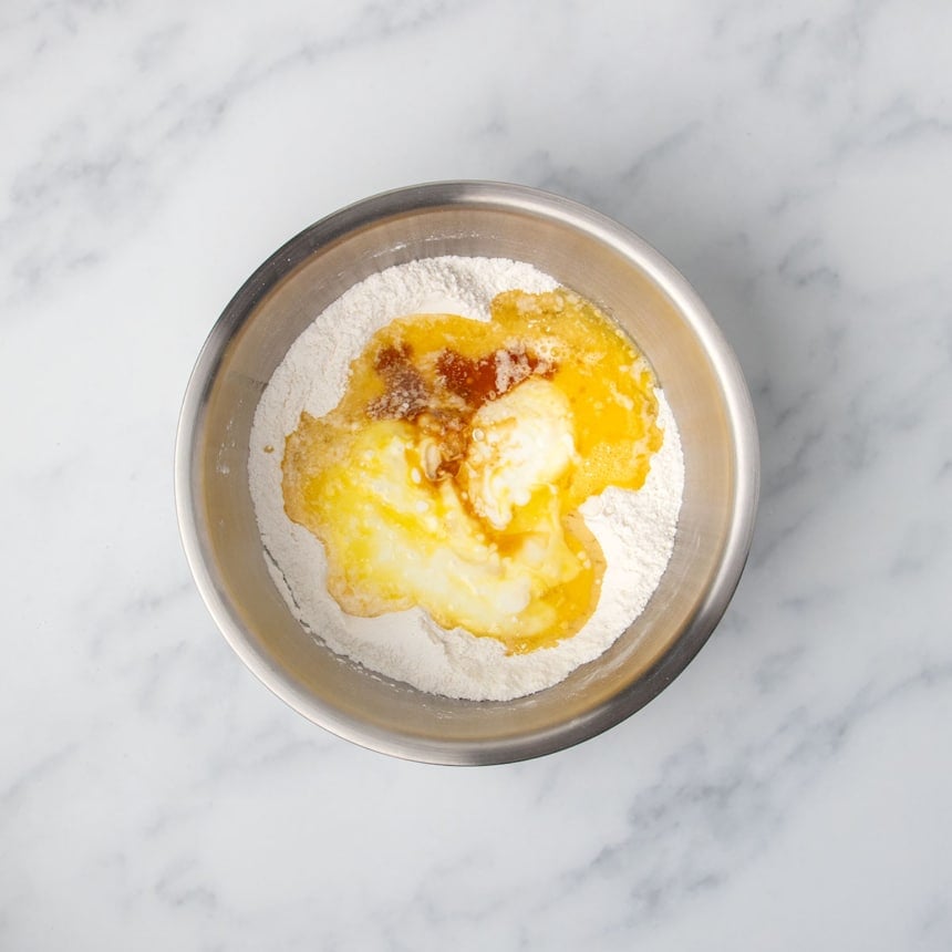 same mixing bowl with dry white powder underneath and white buttermilk, melted yellow butter, oil, brown vanilla extract in center of bowl, unmixed