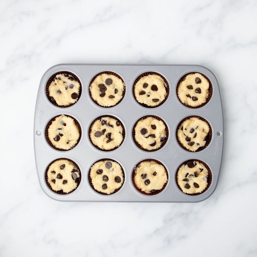 standard 12-cup light colored metal muffin tin with brown paper liners and raw gluten free chocolate chip muffin batter filling each well