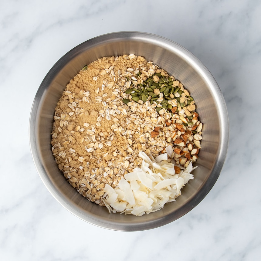 Large round metal mixing bowl with brown oats, sugar and chopped almonds, white coconut chips, and green pumpkin seeds