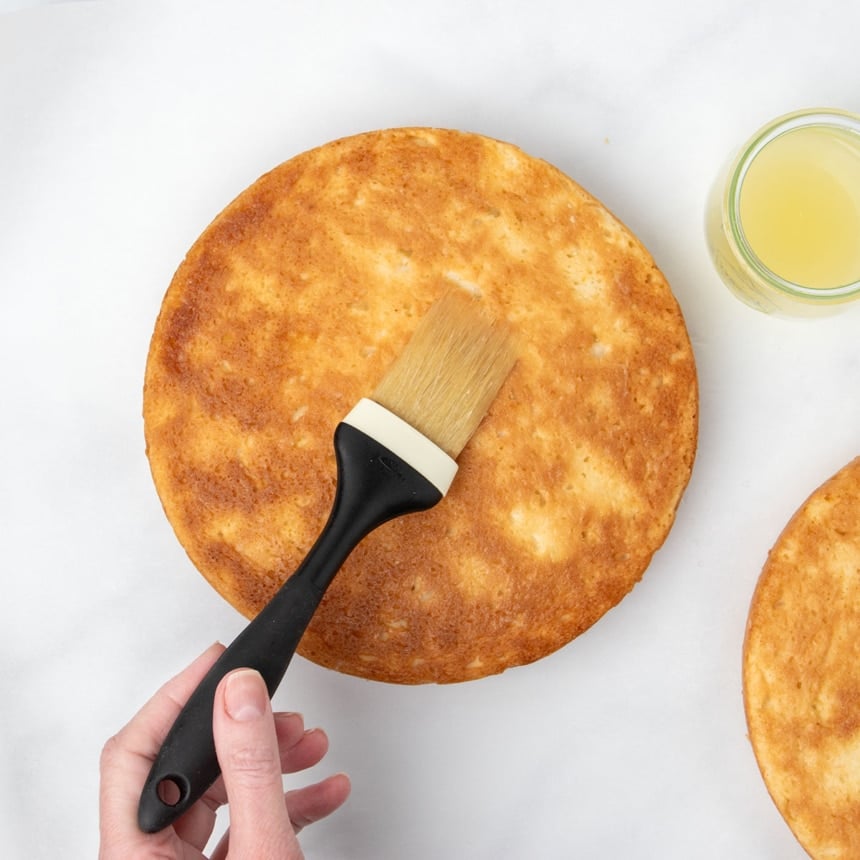 hand holding pastry brush with black handle brushing lemon juice on bottom of baked round light brown and yellow cake, with jar of lemon juice and bottom of second baked cake