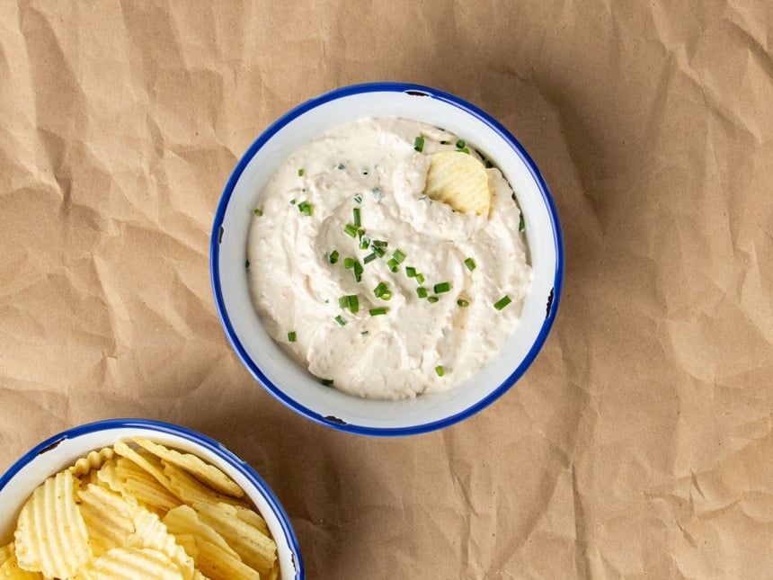 overhead image of onion dip with chopped fresh chives on top with one chip in bowl on brown paper with more chips in another bowl