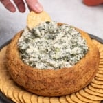 fingers holding round cracker in white and green spinach dip in round brown bread bowl