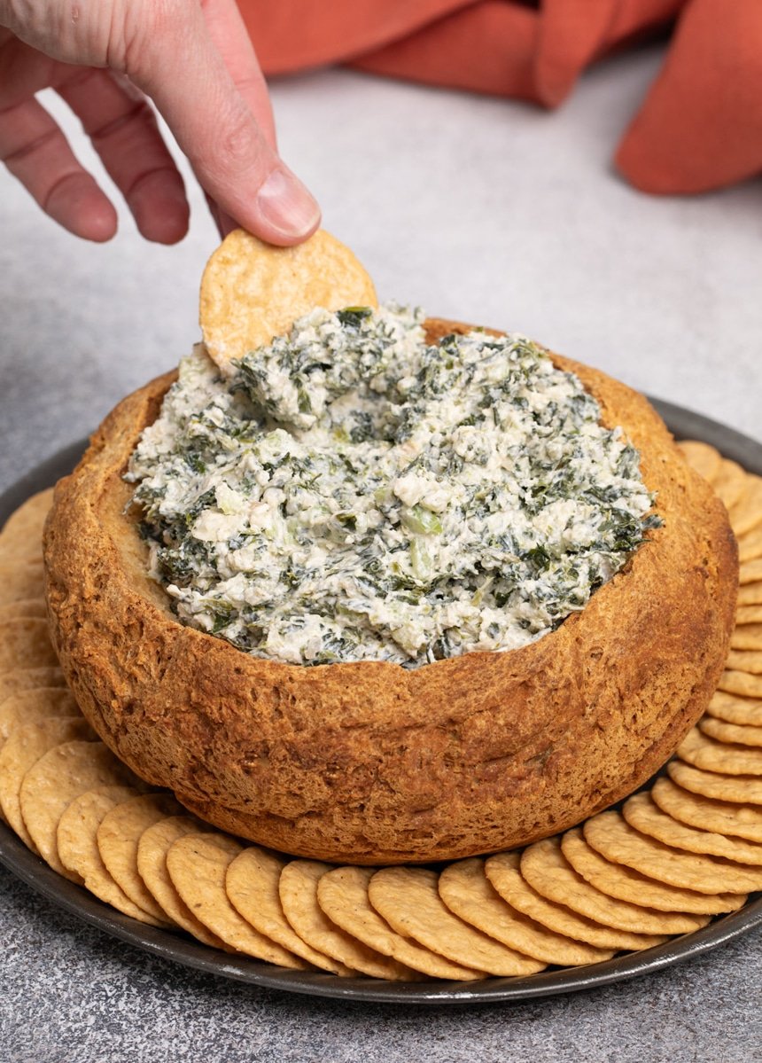 fingers of one hand holding a round light brown cracker scooping thick white and green flecked dip from brown crust of hollowed out bread bowl surrounded by more round crackers