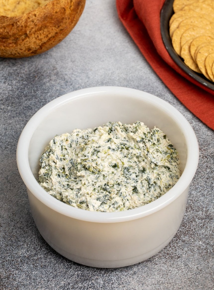 thick white and green flecked spinach dip in round deep white bowl on speckled gray surface with crackers on black plate and bread bowl in background