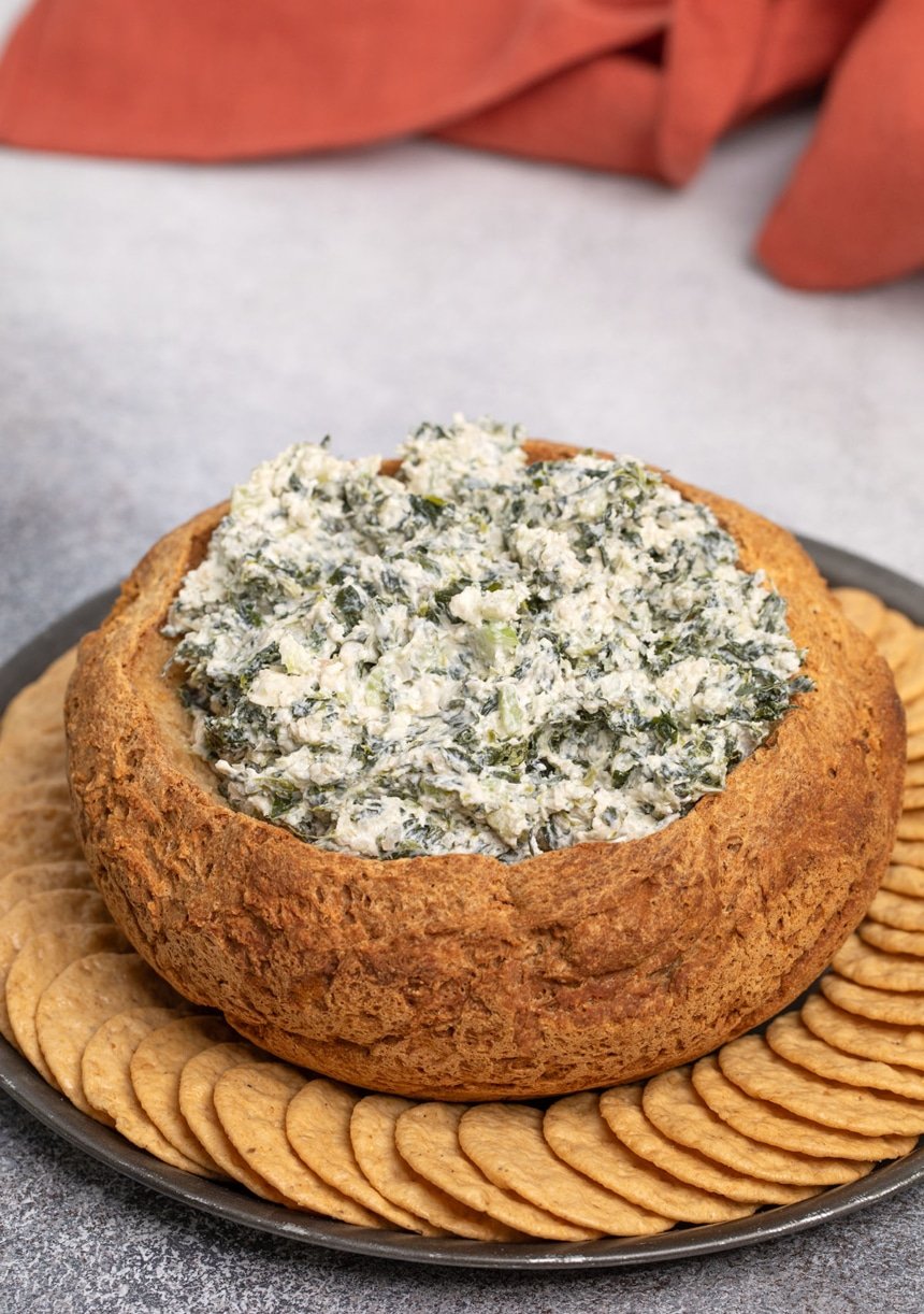 thick white and green dip in brown crusty bread bowl on round black plate surrounded by round light brown crackers