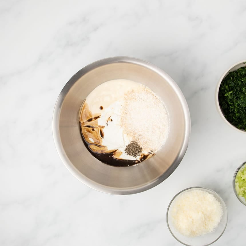 large round metal mixing bowl with white sour cream and mayonnaise, brown Worcestershire sauce, black pepper, and light brown and white powder