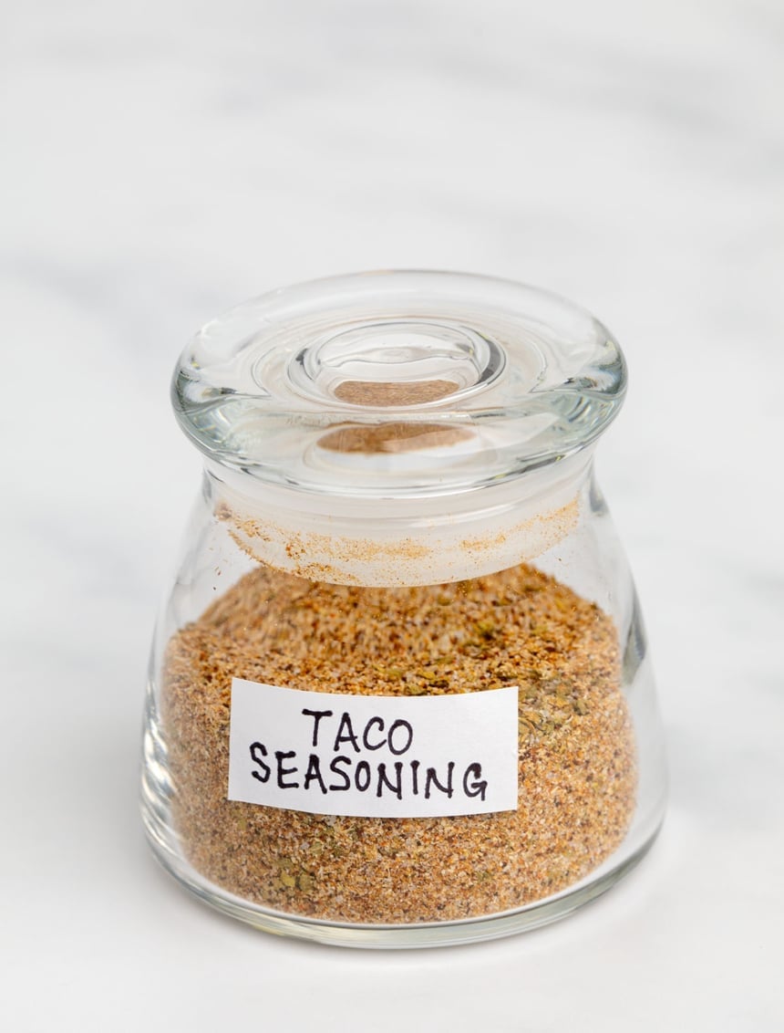 small clear glass bottle with clear lid filled with orange-red-green powder inside and white label with black lettering saying taco seasoning