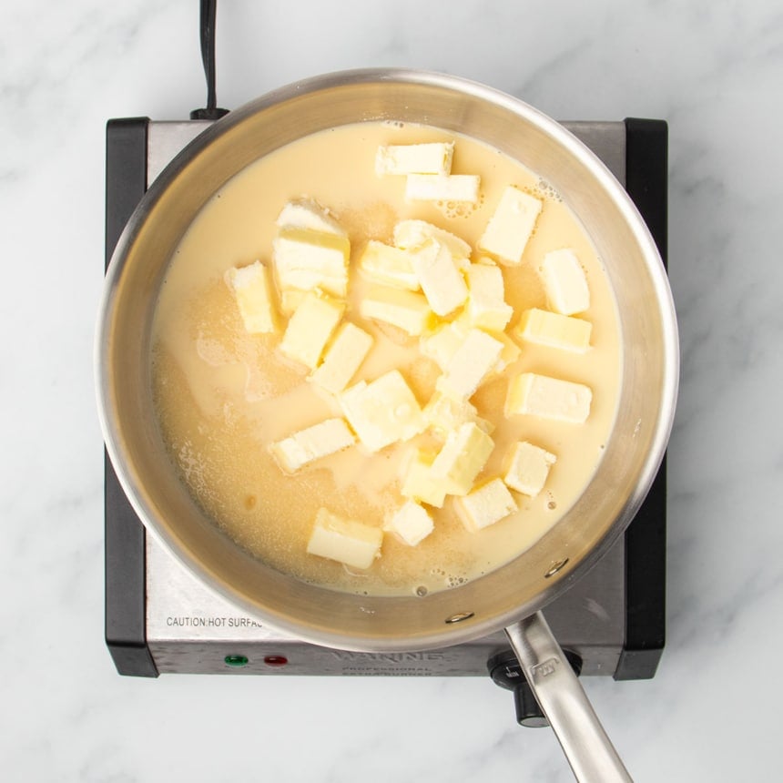 round metal saucier pan with chopped butter pieces, sugar, and light tan evaporated milk on electric burner