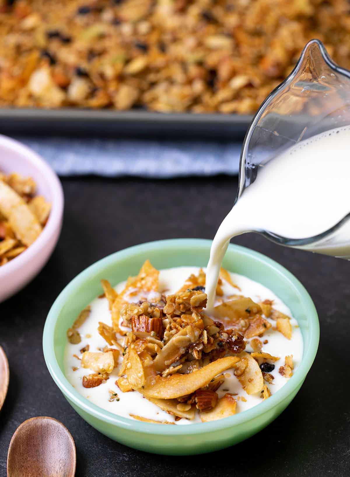 Milk pouring from glass jar into gluten free granola in small green bowl with spoons and pink bowl