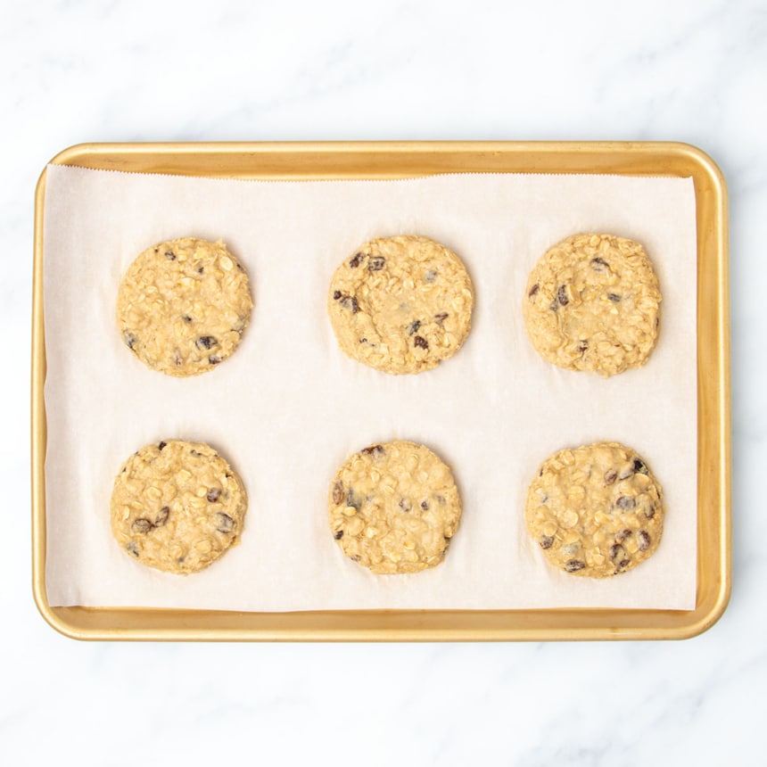 gold colored metal rimmed baking sheet with white parchment paper lining with 6 round raw light brown oatmeal breakfast cookies with dark brown/black raisins