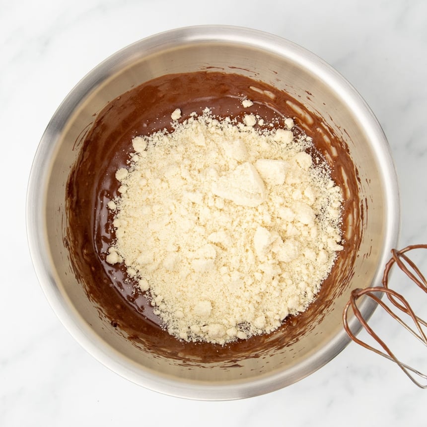 same bowl with brown mixture on bottom and light tan almond flour with some clumps on top, with beaters of handheld mixer with some brown mixture on them visible in the corner