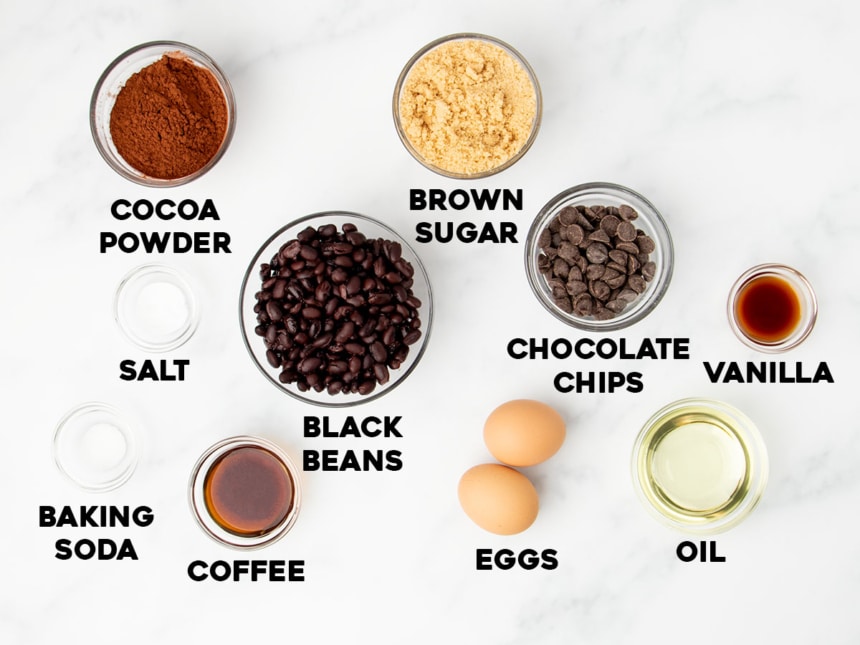 overhead image of ingredients for black bean brownies in small bowls on light marble surface with names of ingredients in black bold type