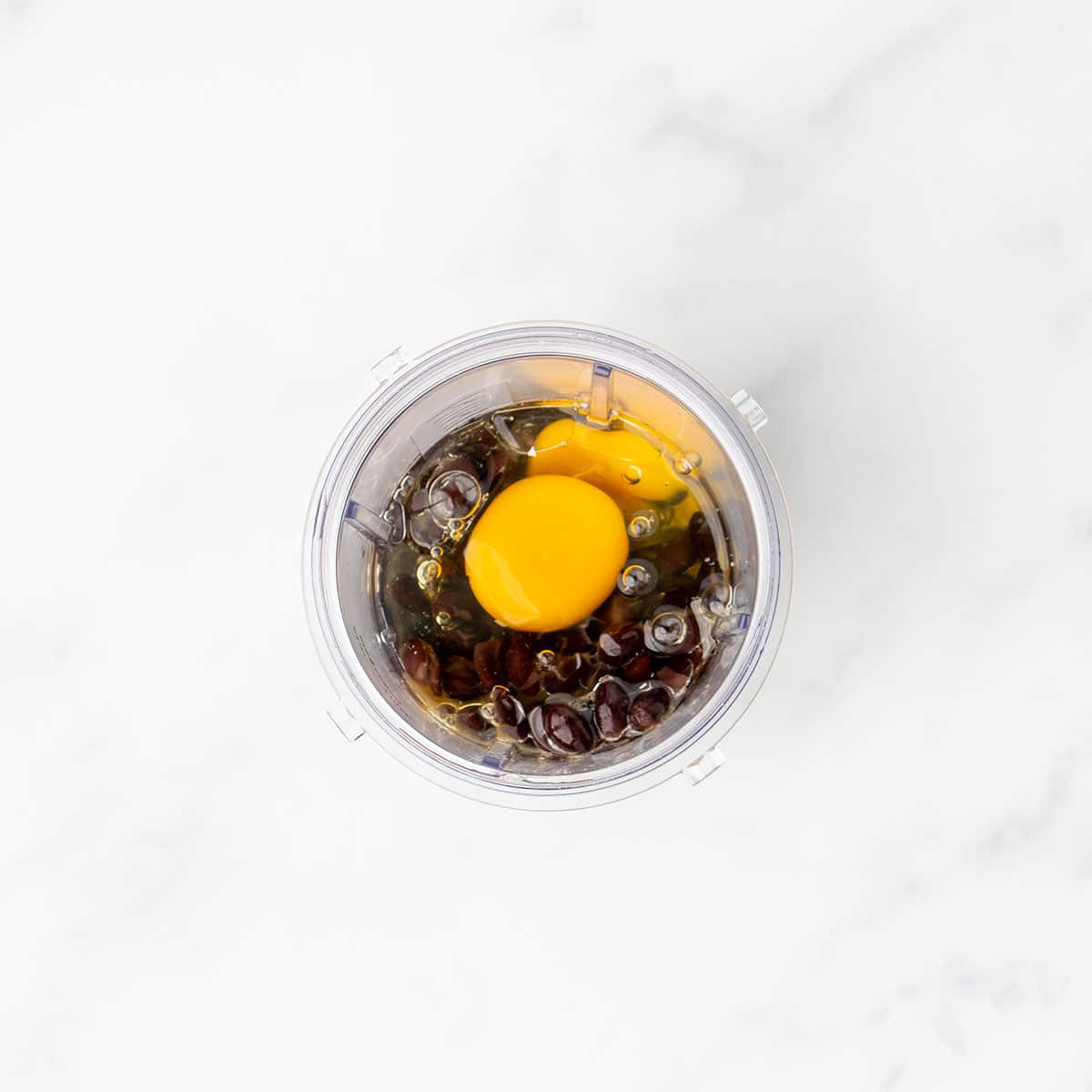Oil, eggs with yellow yolks, vanilla extract and black beans in clear plastic blender canister.