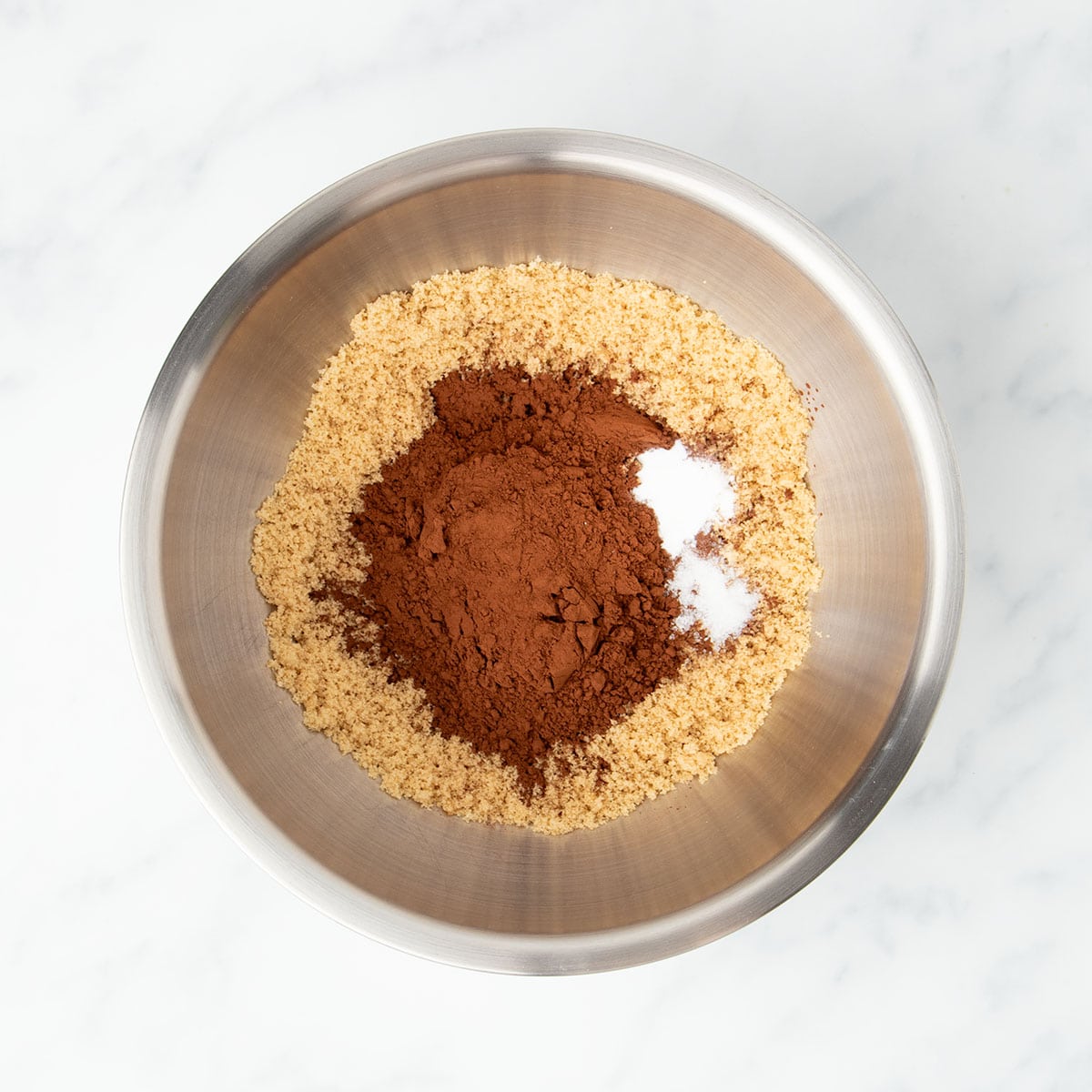 Large round metal mixing bowl with light brown sugar, dark brown cocoa powder, and white baking soda and salt, unmixed.