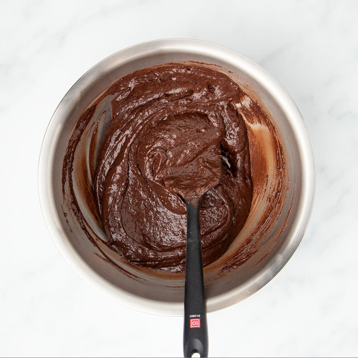Same mixing bowl with soft dark brown raw brownie mixture with black spatula.