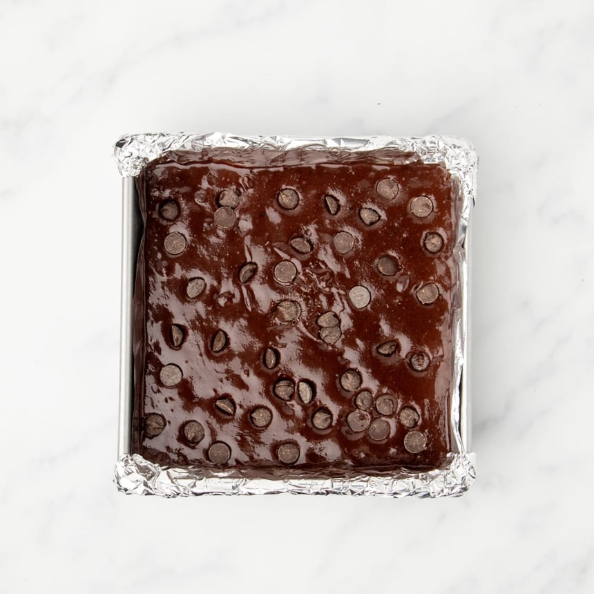 8-inch square metal baking pan lined with foil with dark brown raw brownie mixture with chocolate chips on top