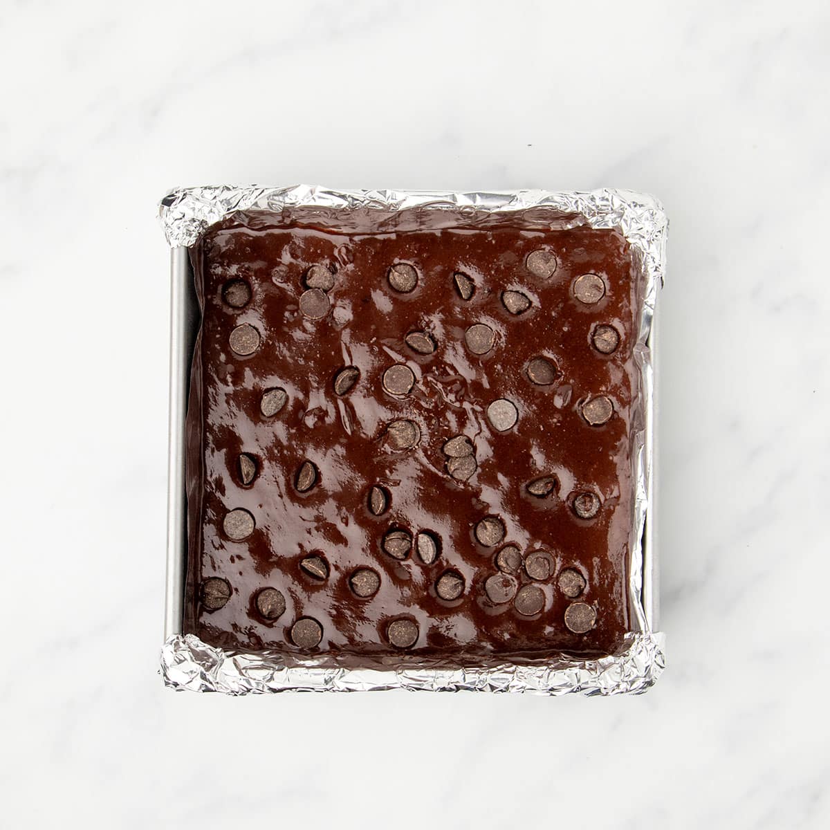 8-inch square metal baking pan lined with foil with dark brown raw brownie mixture with chocolate chips on top.