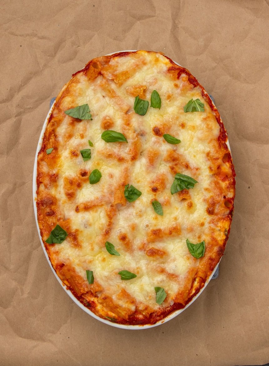 Brown paper underneath whole white oval casserole dish with baked ziti with browned cheese on top and scattered chopped green fresh basil leaves