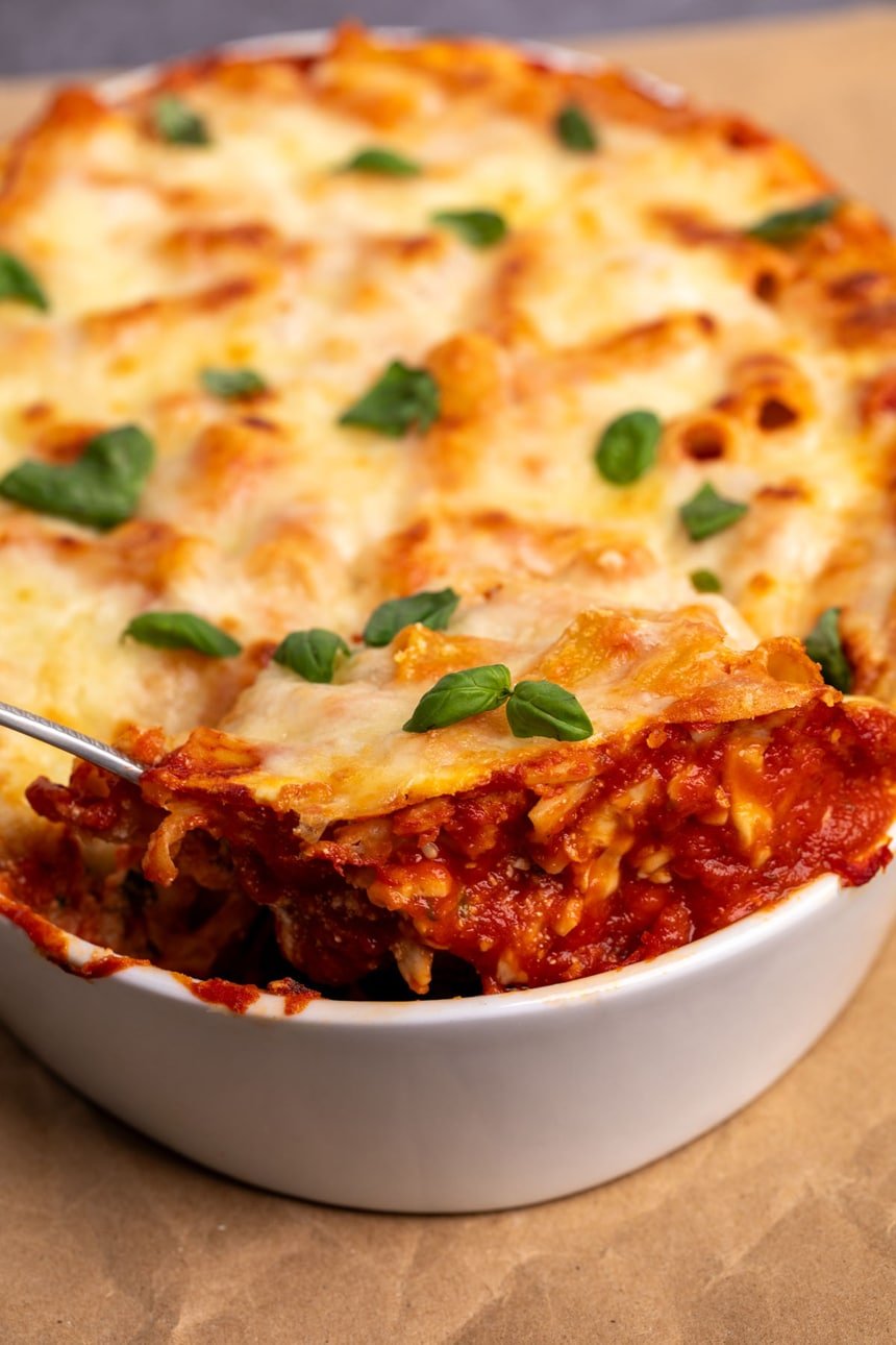 Side of white oval casserole dish with baked ziti being lifted on metal serving spoon revealing tomato sauce, cooked pasta and ricotta cheese, with top layer of browned cheese