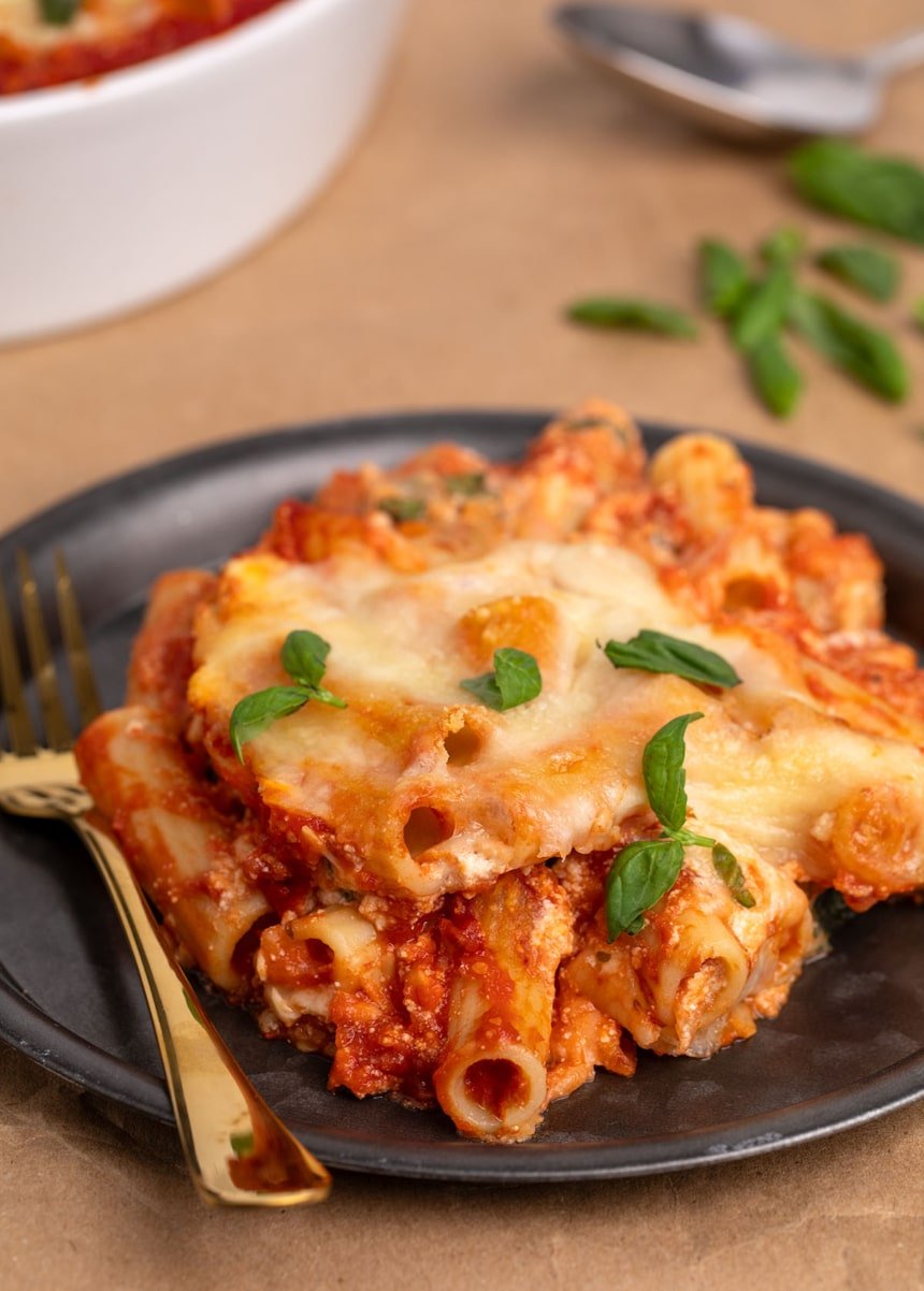 black round dish with gold fork and serving of gluten free baked ziti with ricotta, rigatoni pasta, ricotta and chopped fresh basil from side