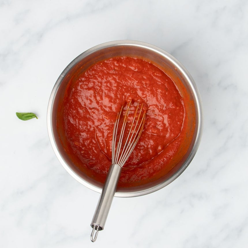 Round metal mixing bowl with red tomato sauce and metal wire whisk in bowl