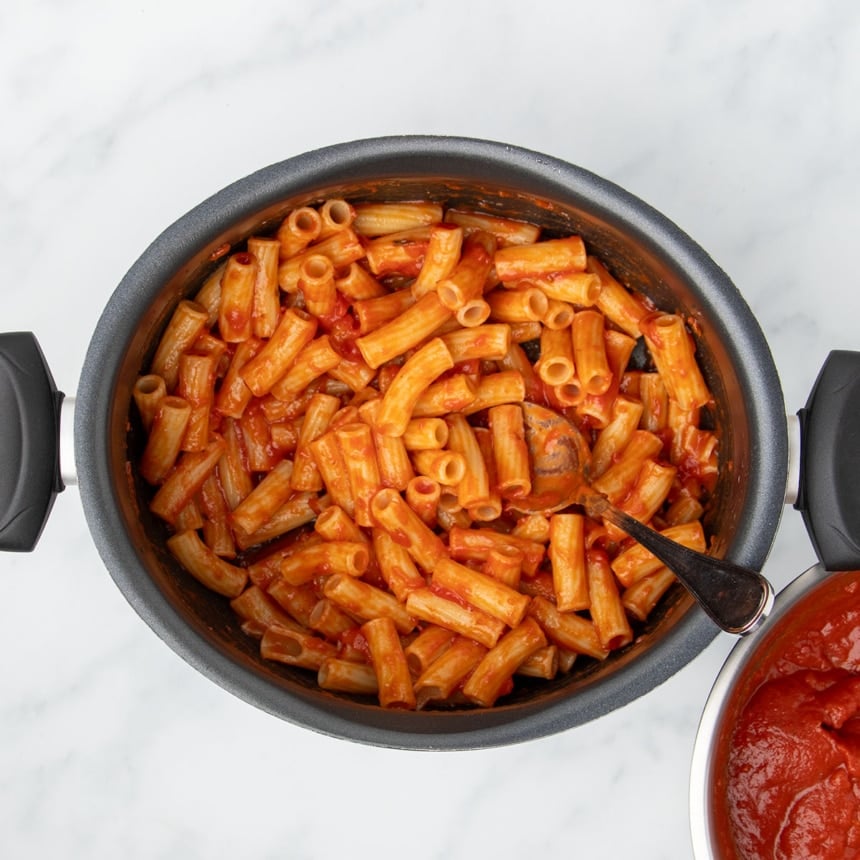 Large black oval stock pot with cooked gf rigatoni noodles covered in tomato sauce with metal mixing spoon