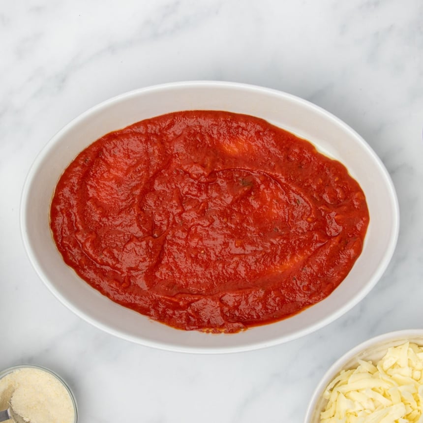 Large white oval casserole dish with layer of red tomato sauce on bottom