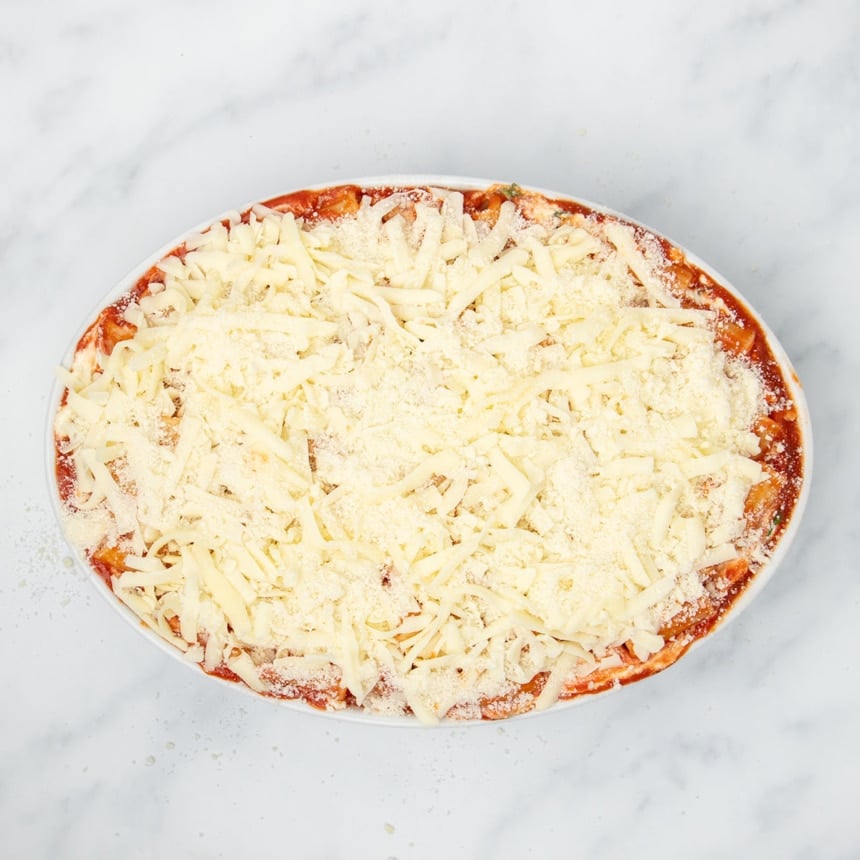 Same dish with top layer of white shredded mozzarella and white finely grated Parmesan cheese with some sauce peeking out around perimeter of dish