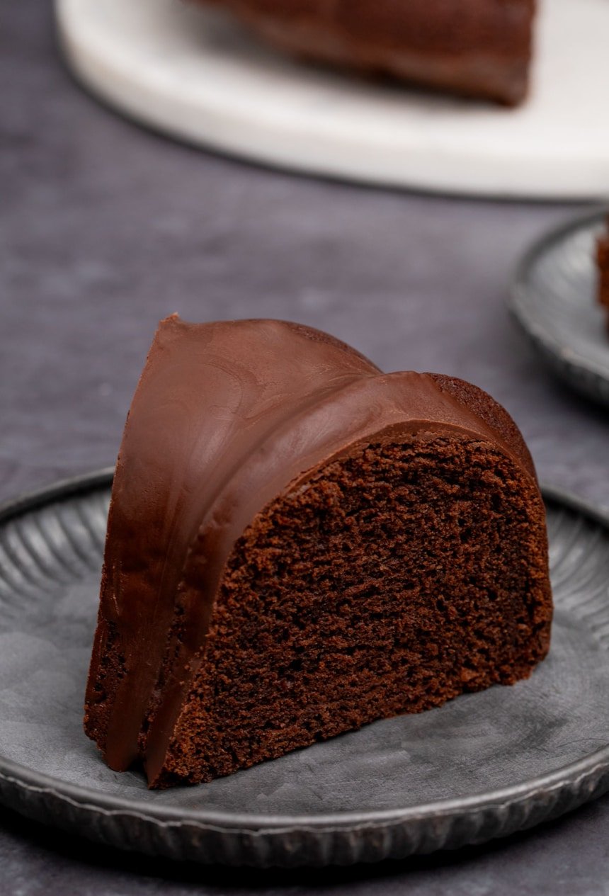 side view of slice of dark brown chocolate bundt cake on dark gray fluted cake plate with rest of cake on white cake plate in background