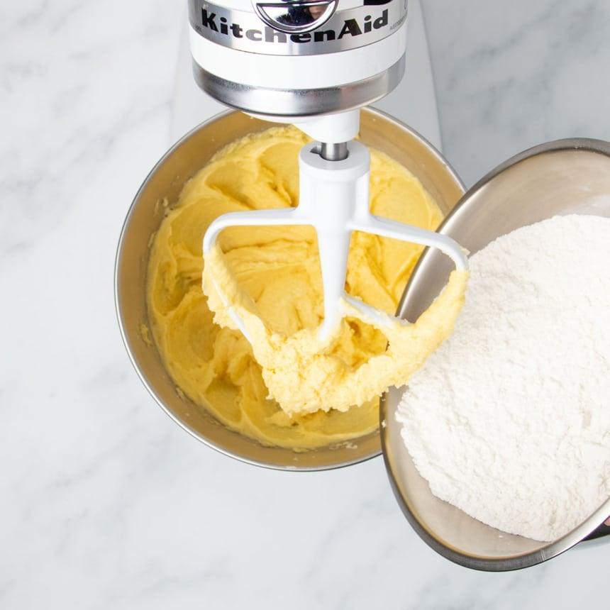 darker yellow mixture in stand mixer bowl with white powder contents of medium bowl being added to the mixing bowl