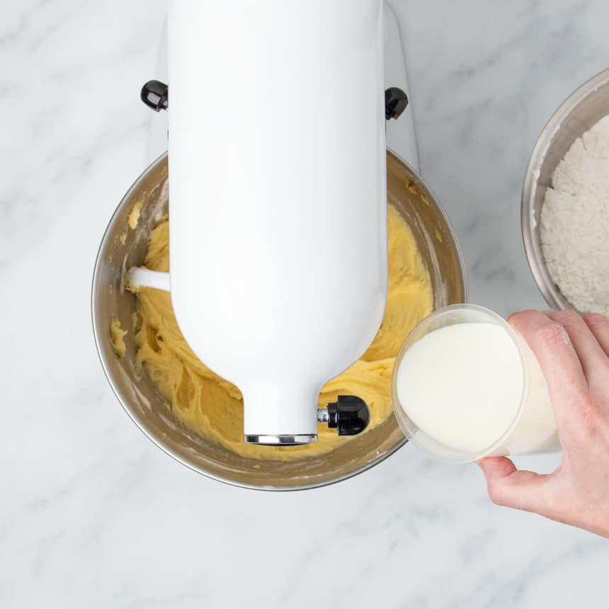 white stand mixer with head tilted down with hand tipping milk in plastic measuring cup into mixing bowl