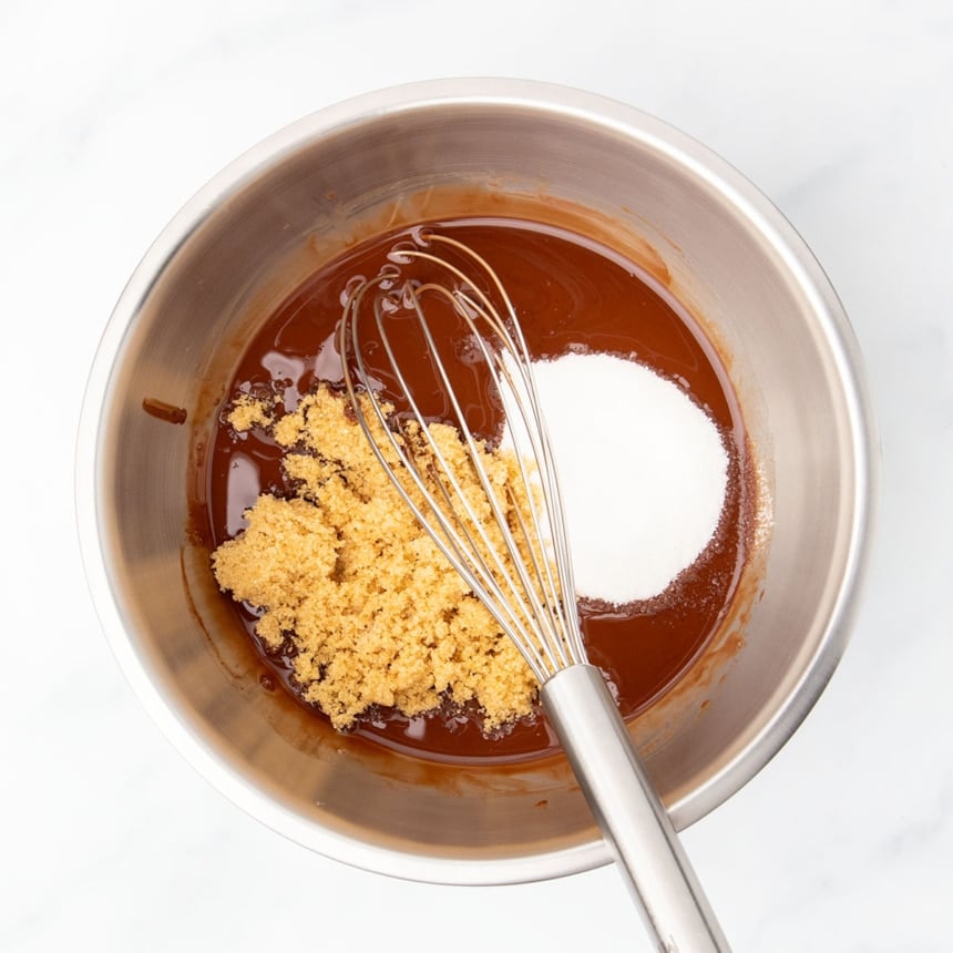 Same medium mixing bowl with whisk with unmixed white sugar and light brown sugar sitting on top