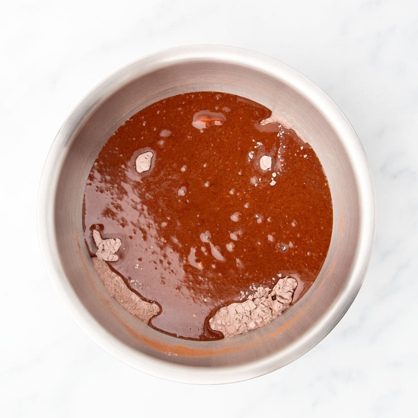 Larger mixing bowl with brown powder on bottom and liquid chocolate mixture poured on top, unmixed