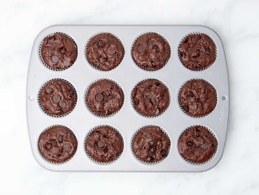 Raw chocolate muffin batter filling all 12 wells of a standard metal muffin tin lined with paper muffin liners