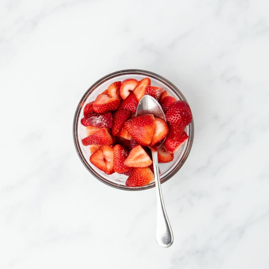 small round glass bowl with cut red and white strawberries and a large metal mixing spoon