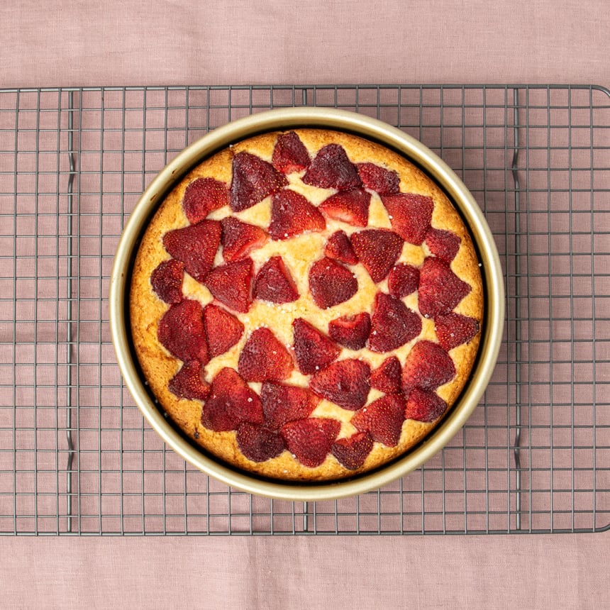 same cake but now baked into a yellow and brown round cake in the same baking pan with darker strawberries on top with a few grains of coarse sugar on a wire metal cooling rack on a light purple cloth