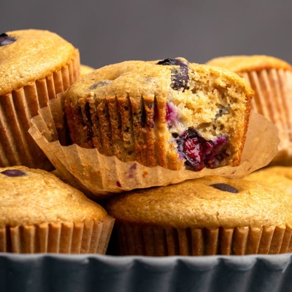 closeup image of healthy blueberry muffin with one bite taken still sitting loosely in brown muffin liner on top of other muffins